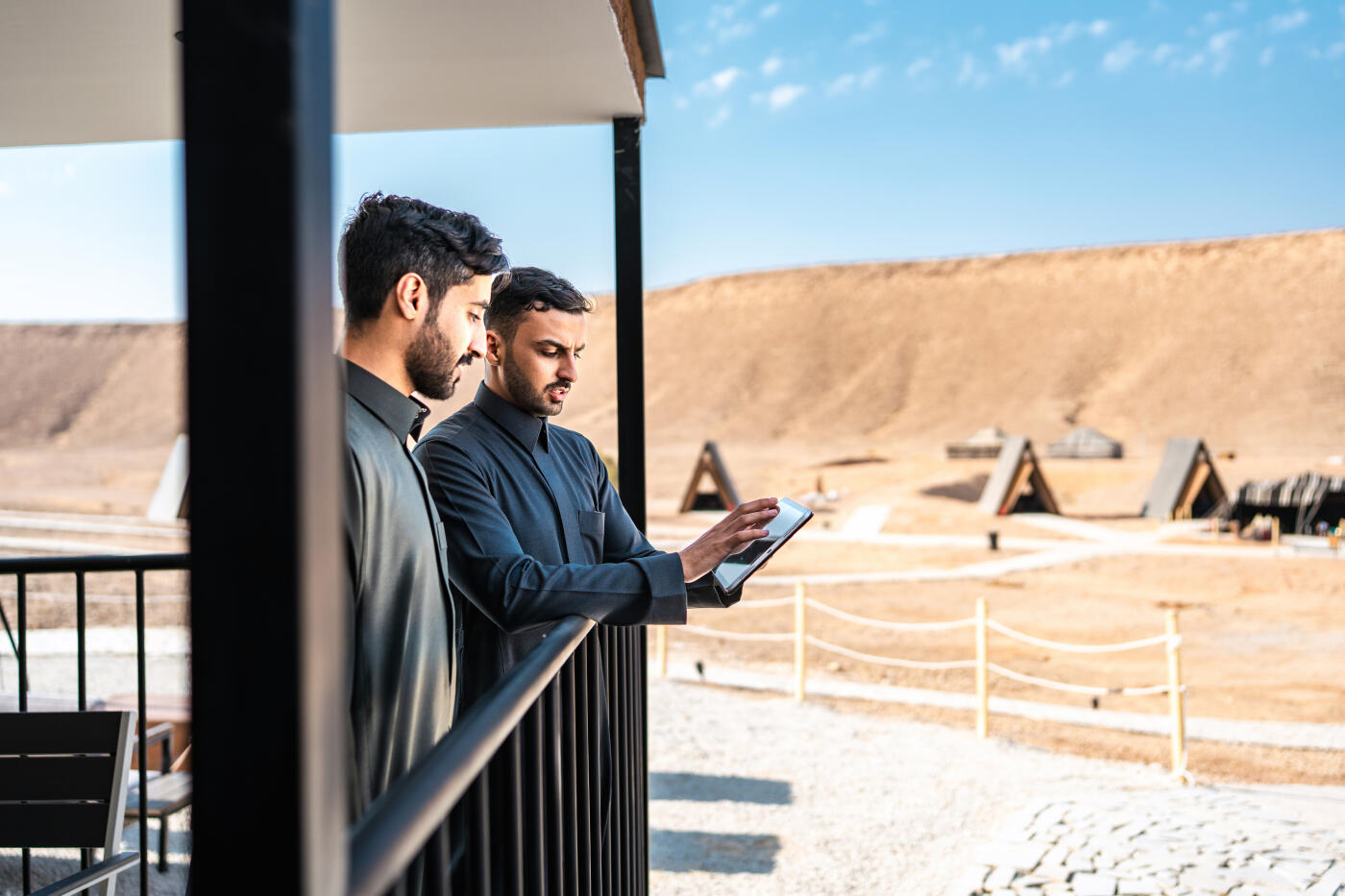 Young middle eastern businessmen collaborating on a digital tablet while standing on the terrace of a business hut at Ghamra Desert Camp. Discussing business strategy and planning. Sunny day with clear blue skies providing natural light.