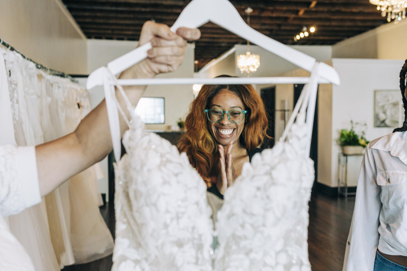 A bride to be smiling excitedly while looking at a delicate and intricate wedding gown being held up by a bridal stylists.