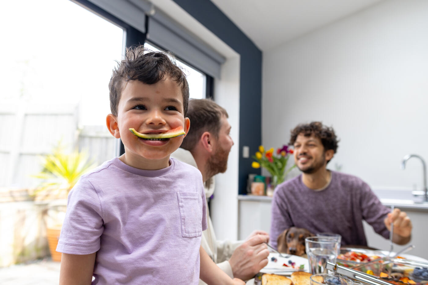 A medium shot of a young boy smiling with the rind of a watermelon slice in his mouth as he looks away from the camera. His two fathers are behind him at the dining table, smiling at each other as they enjoy the homemade food on the table.Videos are available similar to this scenario.