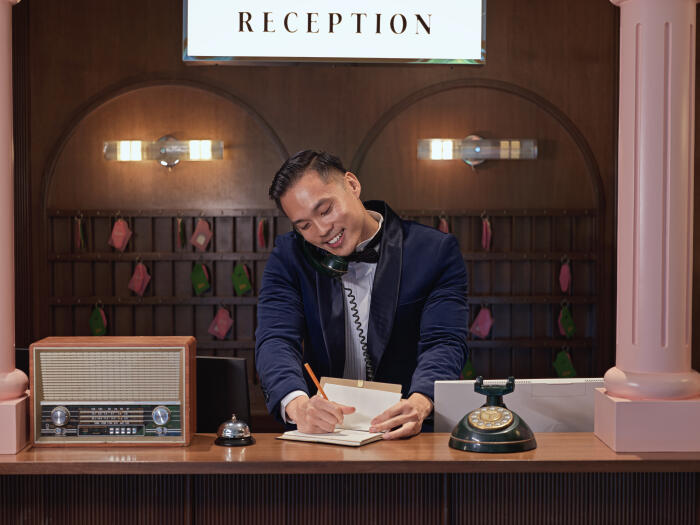 Smiling Asian Chinese male Hotel concierge taking notes while talking on the phone at vintage reception counter, luxury hospitality concept