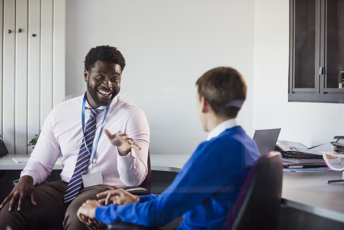 A male secondary school student sitting in an office in the school, engaging in conversation with his teacher. The teacher is smiling and showing positive emotion while pointing as he talks. They are discussing the student's progress.