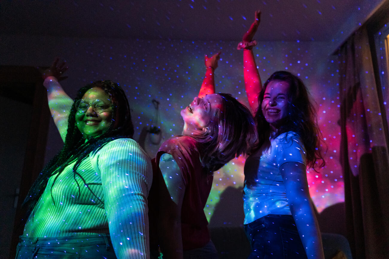 Three cheerful female students are enjoying a fun night together, dancing under the colorful lights of a galaxy projector in their apartment