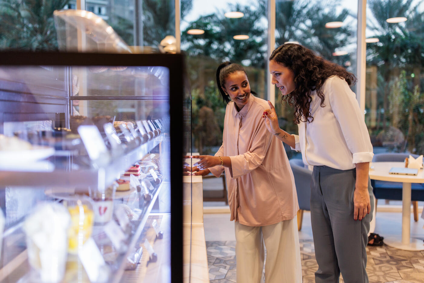 Women smiling and discussing their options while looking at dessert selections inside a display fridge at a stylish coffee shop.