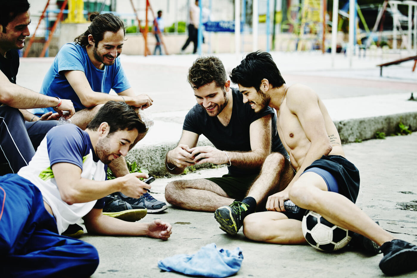 Smiling group of friends looking at smartphones in urban park after soccer match