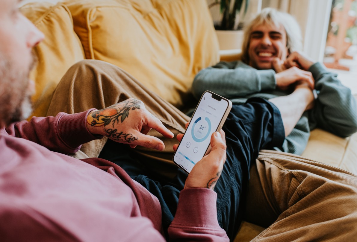 A relaxed gay couple recline on a soft yellow sofa. One man uses a mobile app to control the home thermostat.