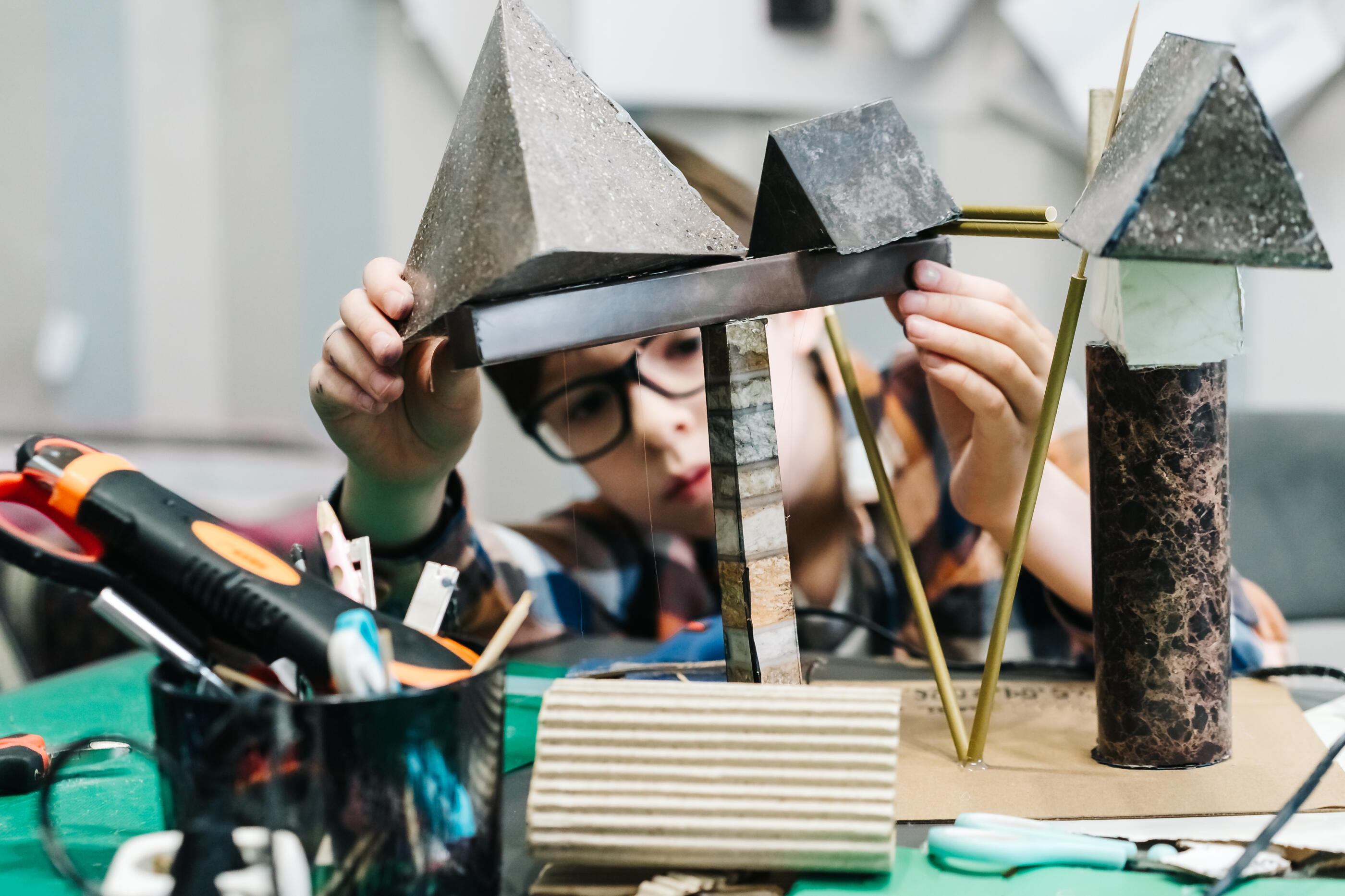 A candid indoor photo shows a child working on a handmade architectural model at a craft table, creating a warm and educational scene focused on creativity and concentration. The image suits themes of learning, DIY projects, childhood imagination, school activities, process of innovation and hands-on making.