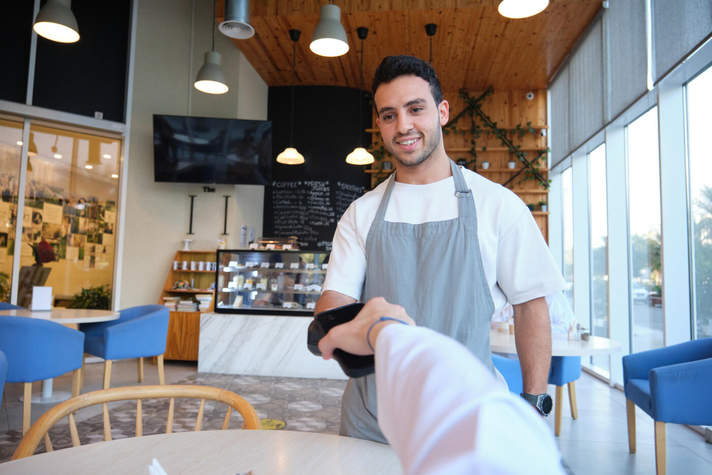 Smiling waiter taking payment from customer at coffee shop. Man is making online transaction through mobile phone. Barista is wearing apron