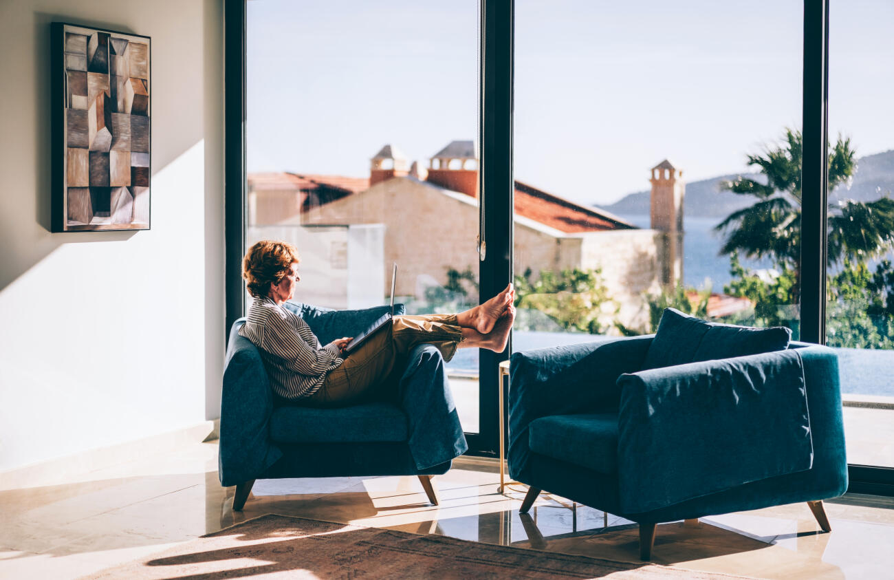 An older woman works on a laptop while sitting comfortably in a stylish living room with floor-to-ceiling windows and a scenic Mediterranean backdrop, a luxury living. The relaxed, sunlit atmosphere blends elegance and technology, ideal for illustrating themes of senior independence, remote living, longevity, or post-retirement digital engagement.