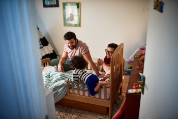 Two fathers watching toddler son playing in bed