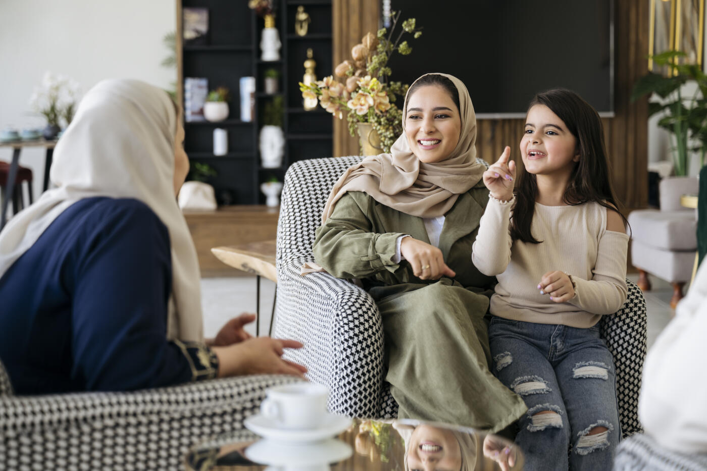 Family gathering including three generations of women in traditional Saudi and western attire smiling and enjoying each other’s company in living room.