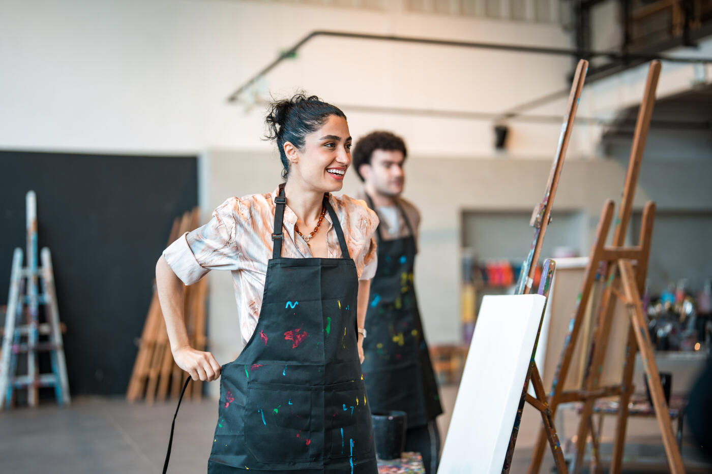 A young Middle Eastern woman wearing a paint-splattered apron prepares for an art class in a spacious, modern studio. A male classmate stands in the background near easels and art tools.