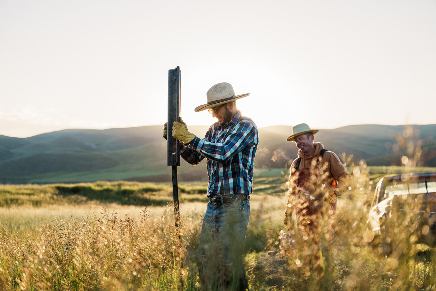 At dawn, two ranchers install a new fence post for an electric fence in a hay field, illuminated by the warm morning light on their Wyoming ranch.