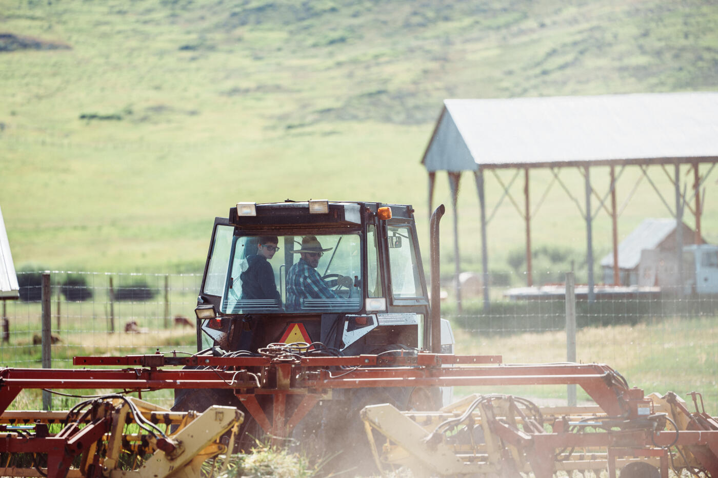 A tween boy rides in a tractor while learning from his dad how to operate the equipment as it tows a rotary rake to prepare cut hay for baling during harvest season on their Wyoming ranch.
