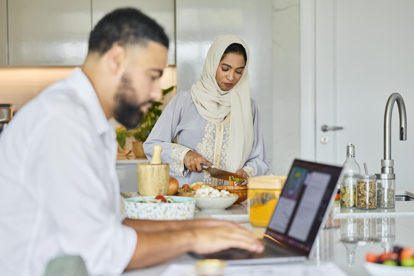 Middle eastern woman cutting vegetables in kitchen. Emirati female in abaya with husband at home. He is using laptop.