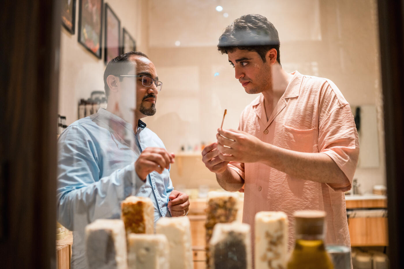 Two men, one in a light blue shirt and the other in a peach shirt, examine traditional handmade soap in an indoor Al Balad shop setting.