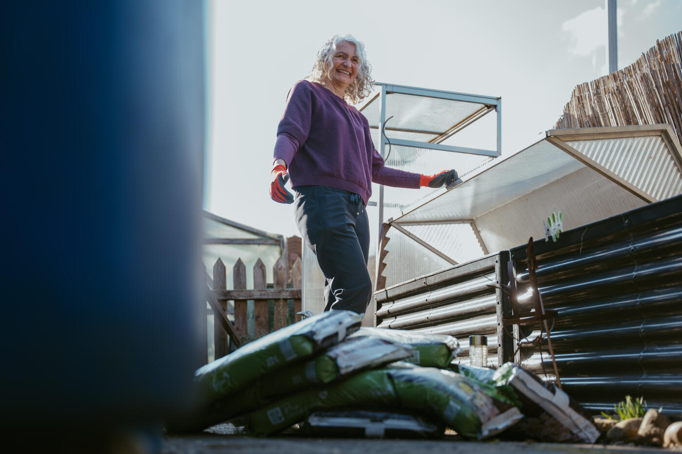 Middle-aged woman wearing gardening gloves, standing next to a raised garden bed and smiling while working. Several bags of soil are placed on the ground nearby. The image represents home gardening, preparation for planting, and a positive outdoor lifestyle