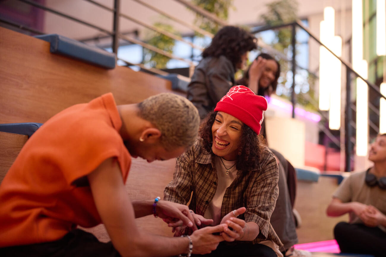 Male and female friends laughing while engaging in conversation. Group of young entrepreneurs talking in shared seating area. Concept of workplace culture and friendship.