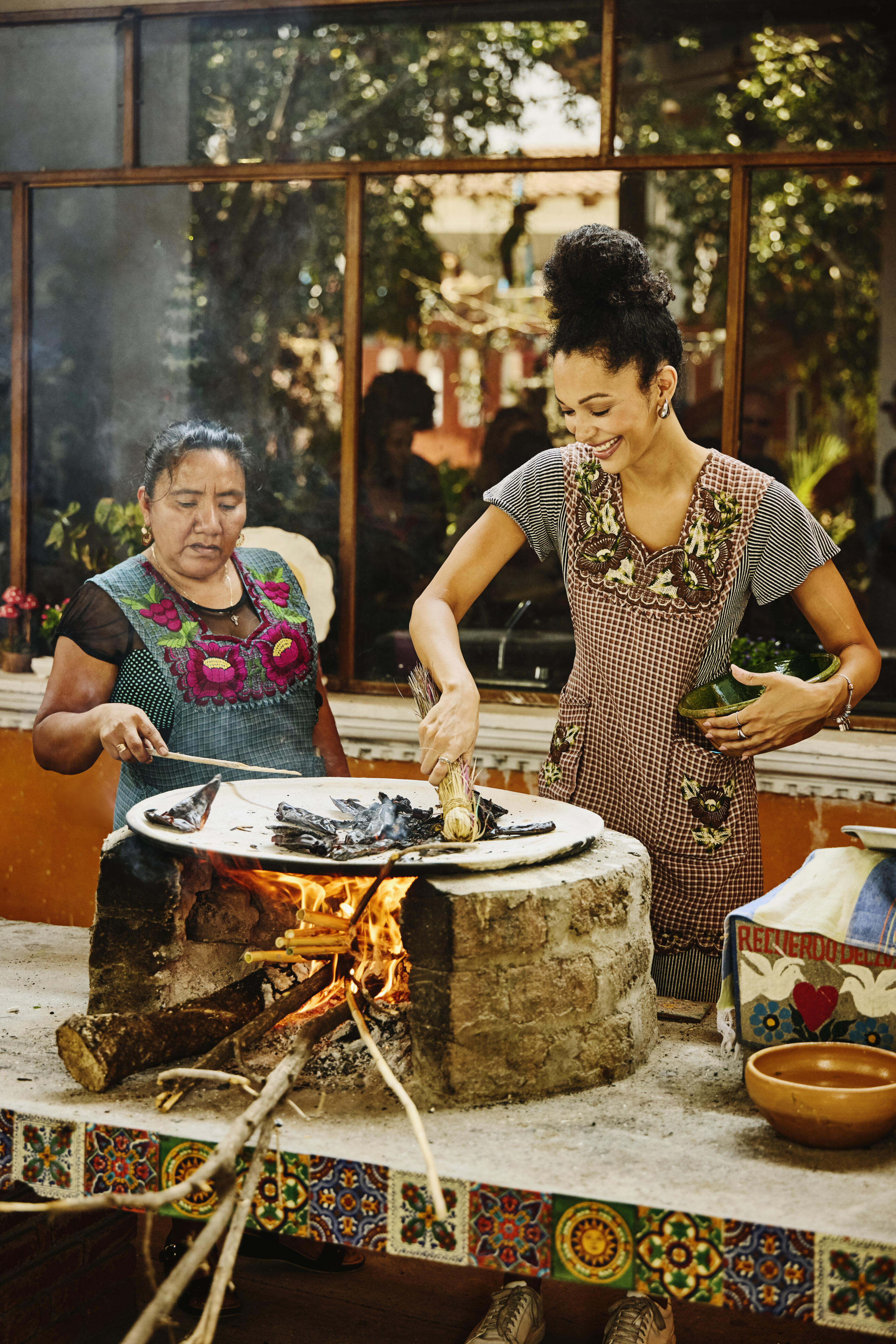 Medium wide shot of smiling female student roasting chili peppers on traditional comal grill during Mexican culinary workshop