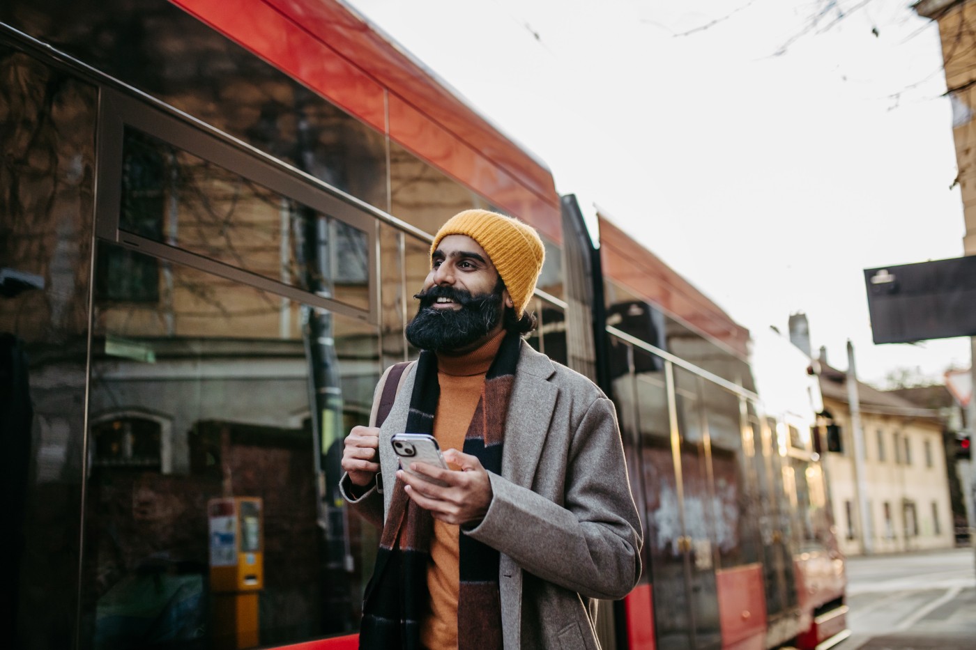 Young Indian man walking through the city during cold spring day. Portrait of handsome indian man in coat, holding smartphone, smiling.