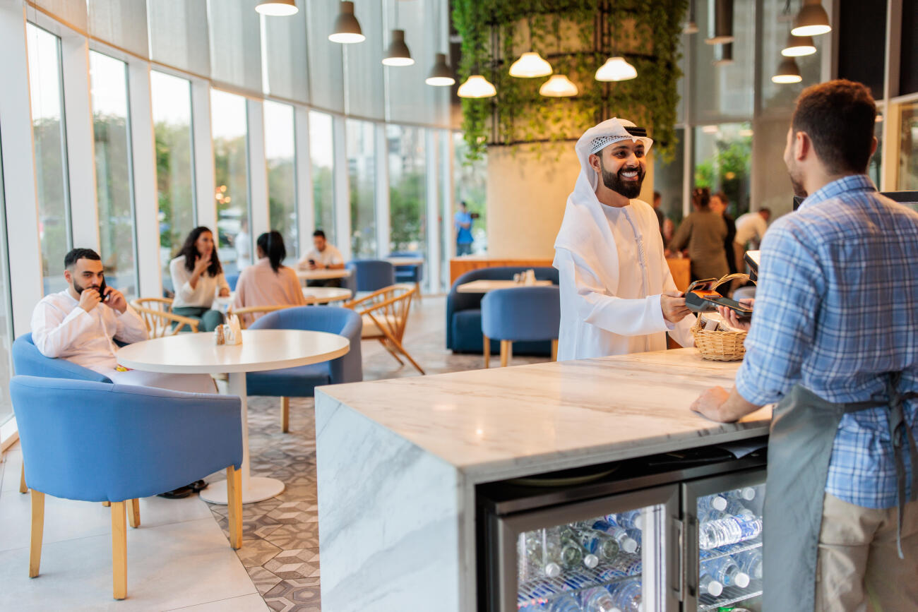 Emirati man in traditional clothing completes a purchase using mobile payment at a café in Dubai.