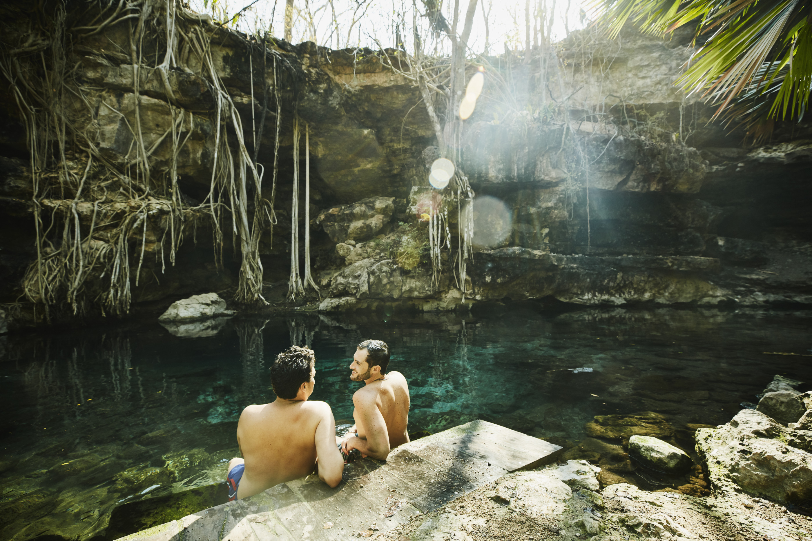 Smiling gay couple in discussion while sitting on edge of cenote after swim