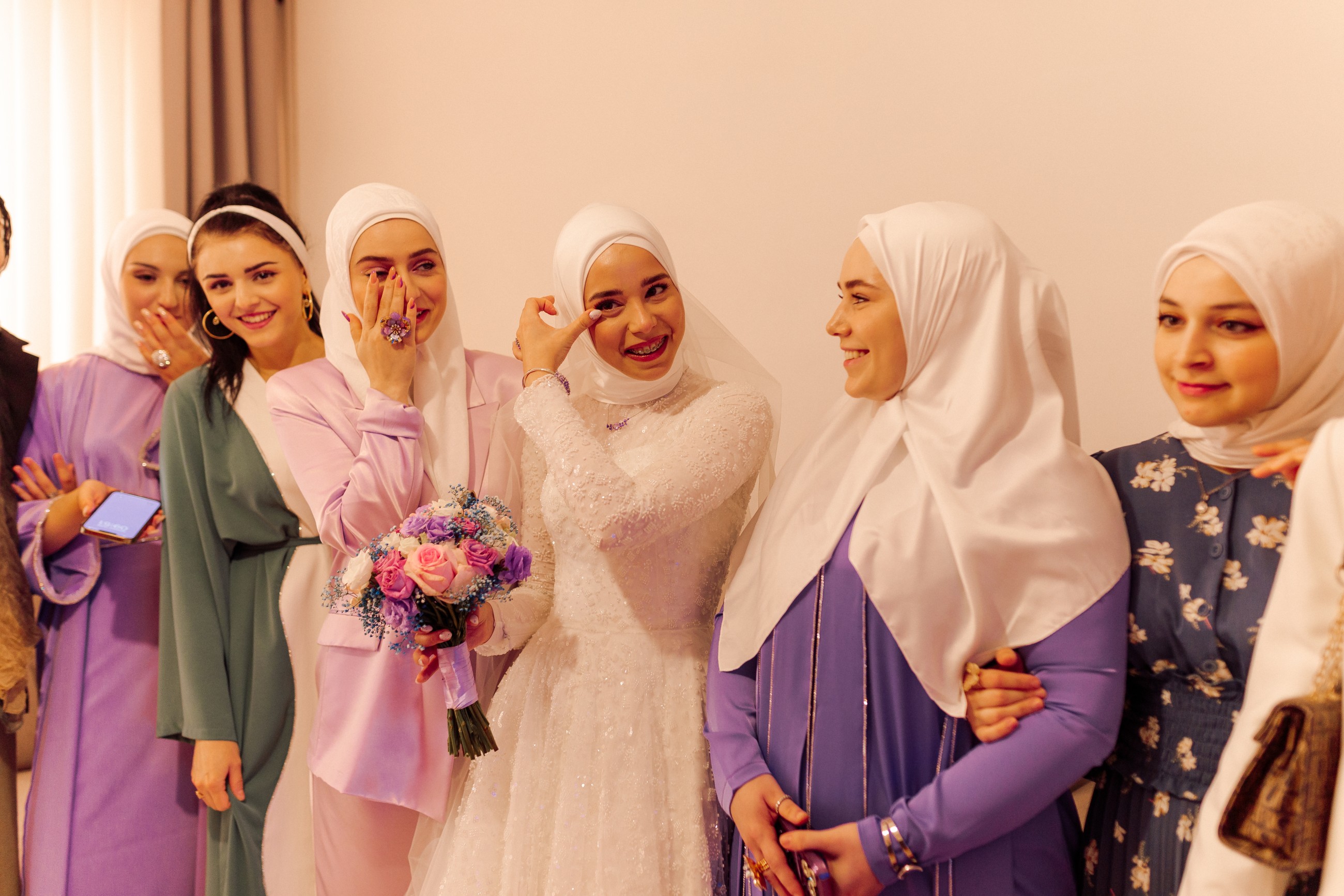 Cheerful young bride in modest white dress and headscarf with veil lines up with delighted female guests