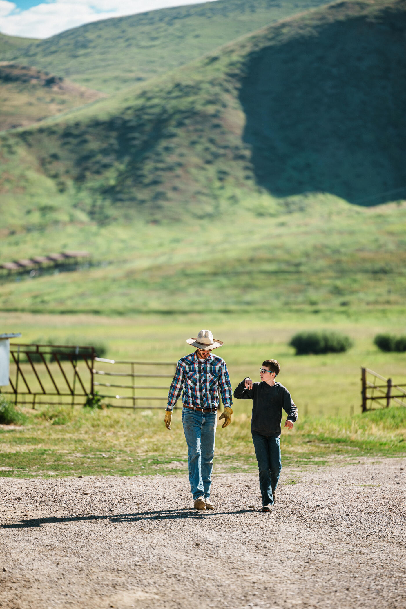 A rancher and his tween son chat as they walk together down a gravel road on their Wyoming ranch on a sunny afternoon.