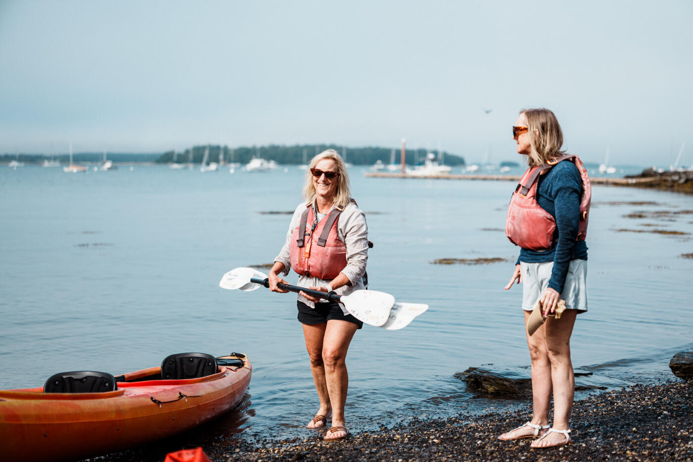 Two smiling senior adult sisters wearing lifejackets and holding oars stand on the shore of a beach in Portland, Maine and prepare to kayak in Casco Bay, visible in the background where sailboats are moored.