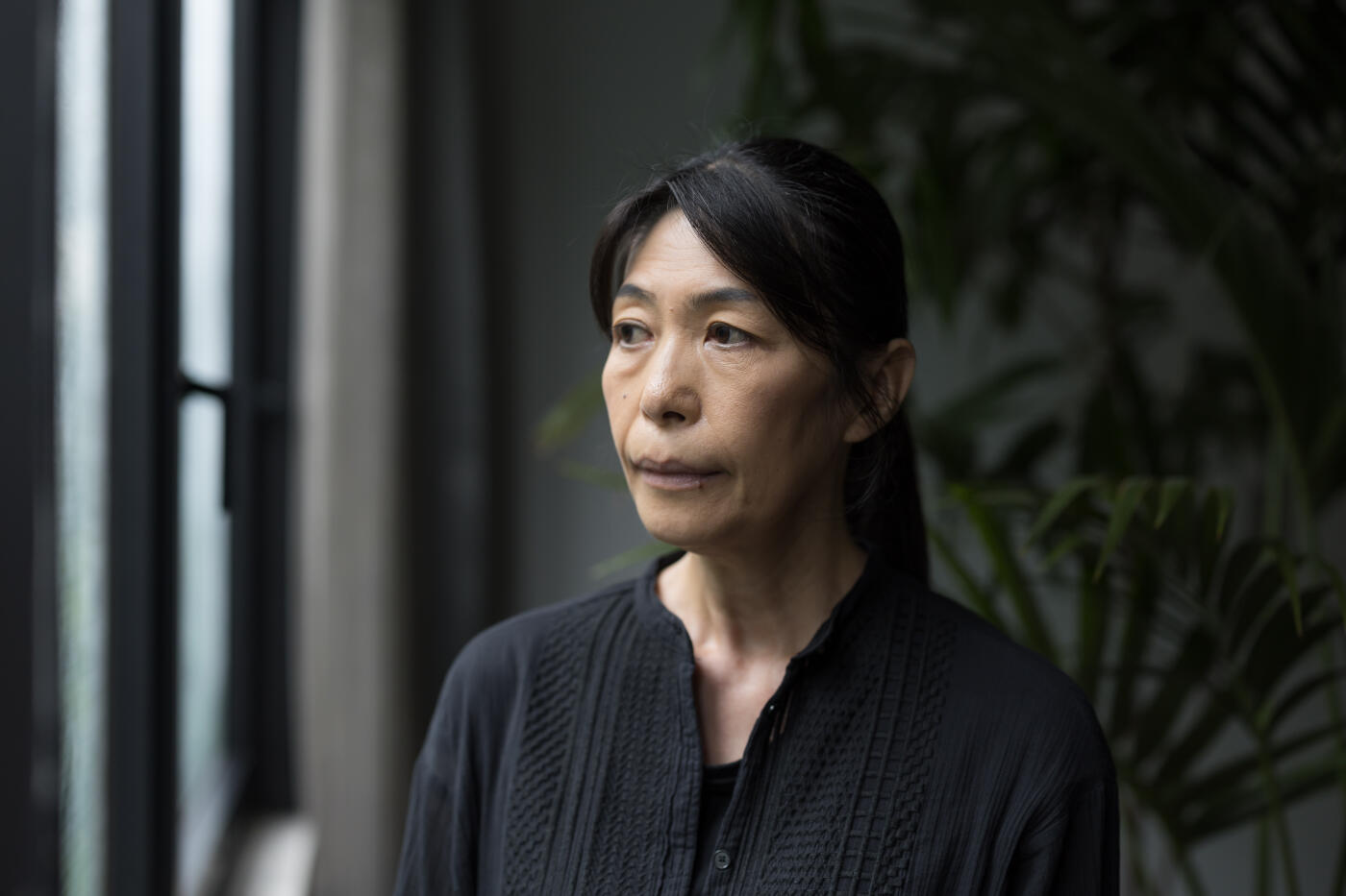 A woman standing by an indoor window looking out the window.