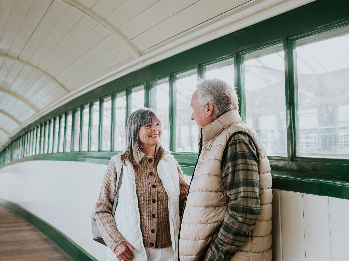 An elderly couple shares a warm moment together while standing by a window in a covered walkway. The woman smiles brightly at the man, creating a joyful and affectionate atmosphere. She wears a knitted sweater with a white quilted vest, while he is dressed in a beige padded vest over a plaid shirt. Natural light filters through the windows, highlighting their expressions and capturing a sense of love, companions