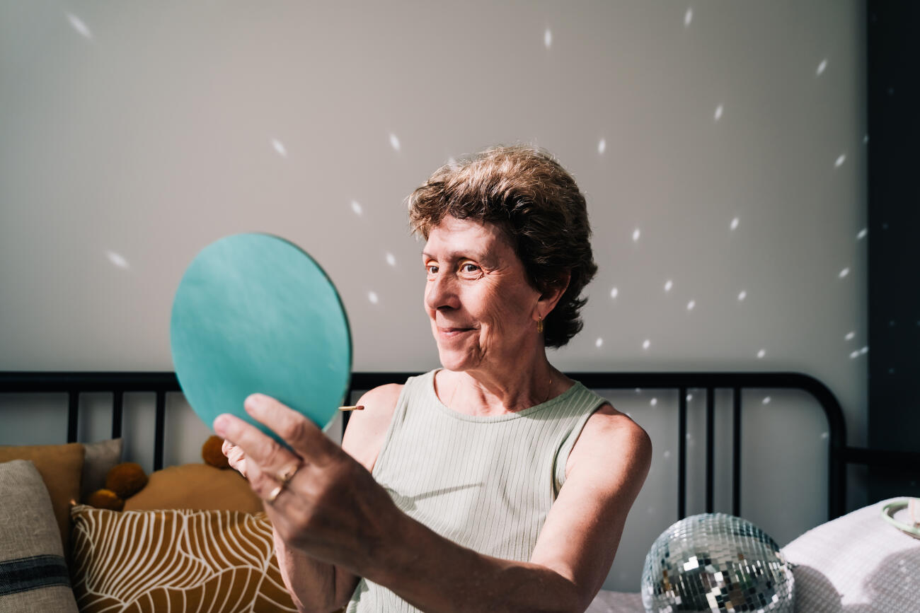A cheerful older woman holds a turquoise mirror and looks at her reflection with curiosity and joy.