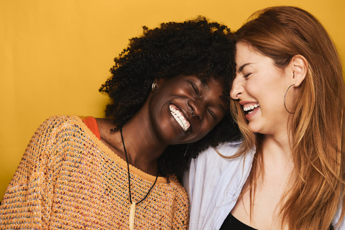 Two happy young adult women with contrasting features sharing a moment of genuine laughter and togetherness, celebrating friendship against a vibrant yellow background