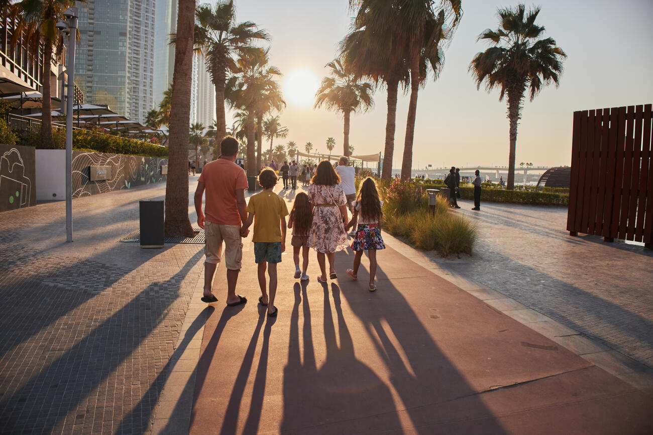 Family enjoying sunset walk along a beach promenade with palm trees and cityscape