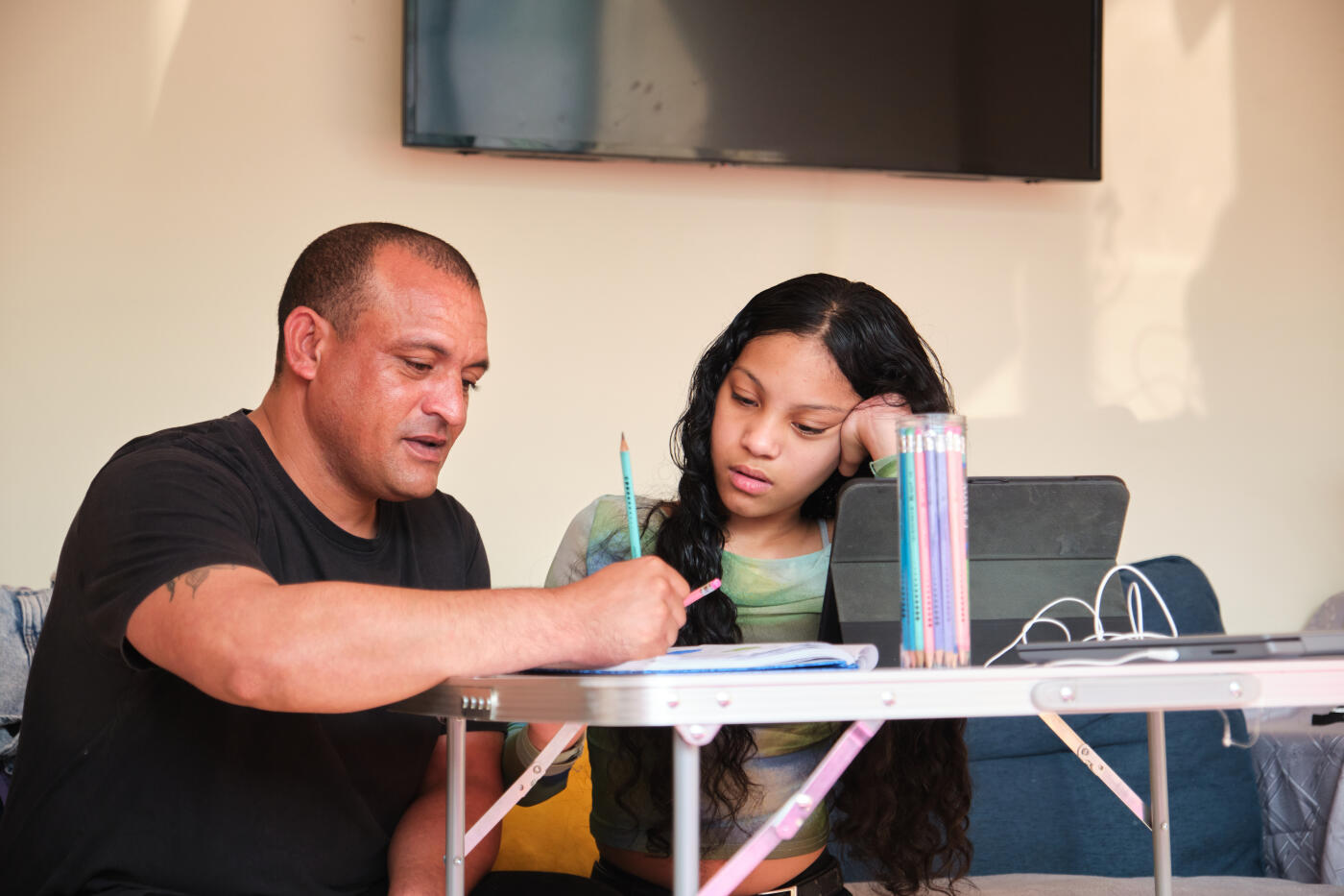 Man writing in book sitting next to daughter while studying at home. Girl is listening to her father explaining homework. They are sitting on sofa.