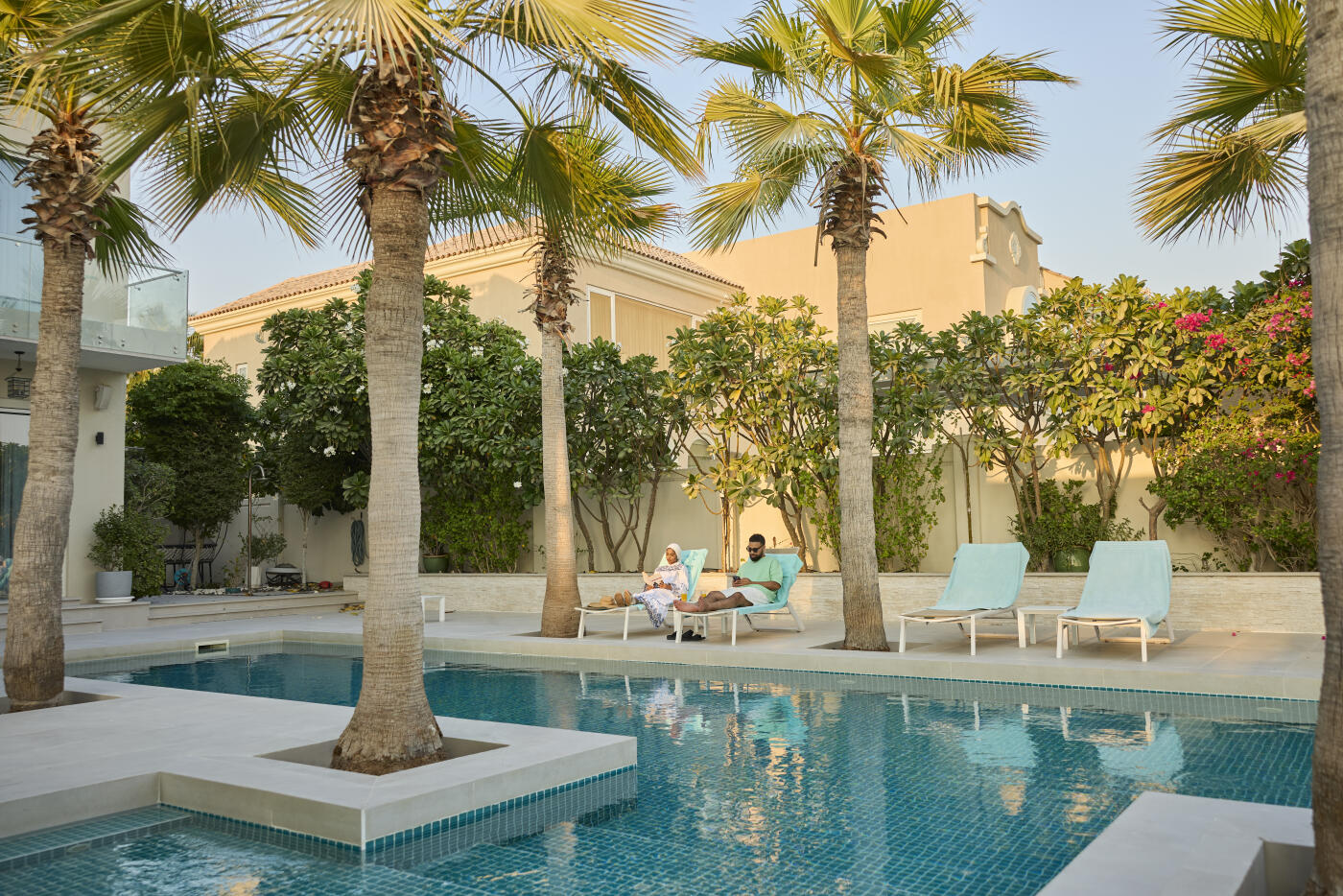 Middle eastern couple spending vacations in luxury villa. Man and woman sitting on deck chair at poolside. Swimming pool can be seen with palm trees
