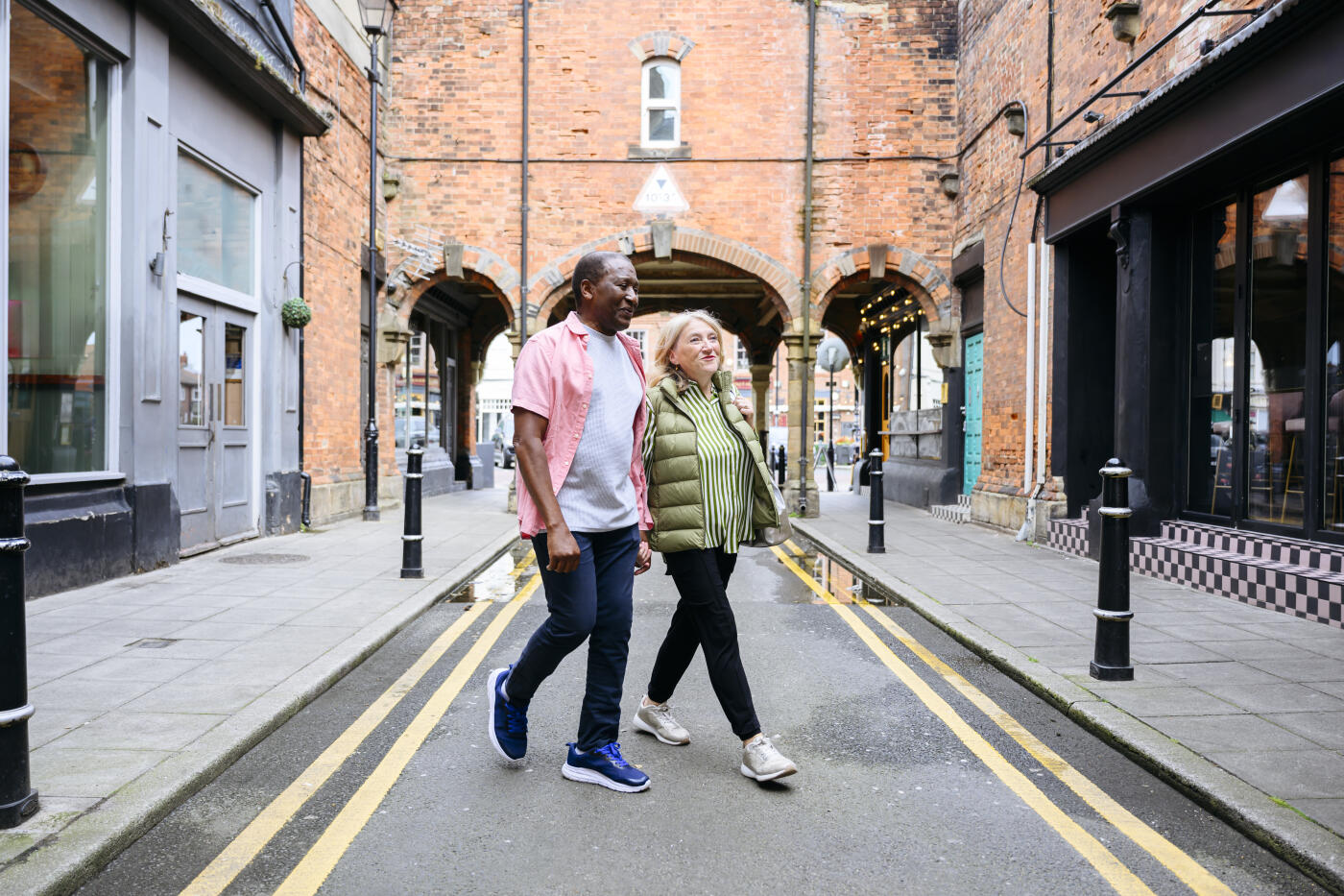 Senior man and woman in casual clothing on narrow road with double yellow lines, walking past independent shops