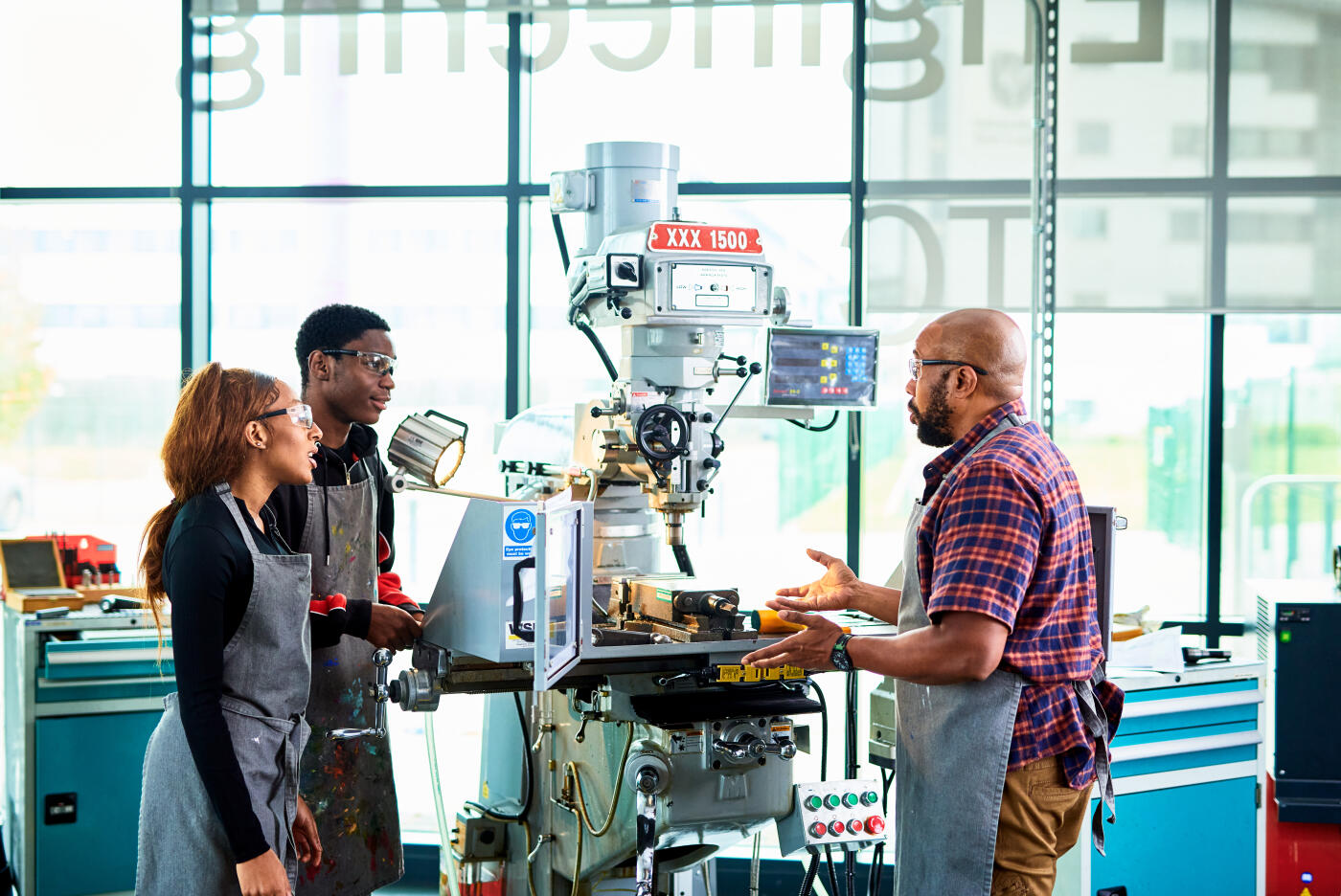 Black mature male teacher talking to two design technology students, explaining and gesturing how to use milling machine in college workshop