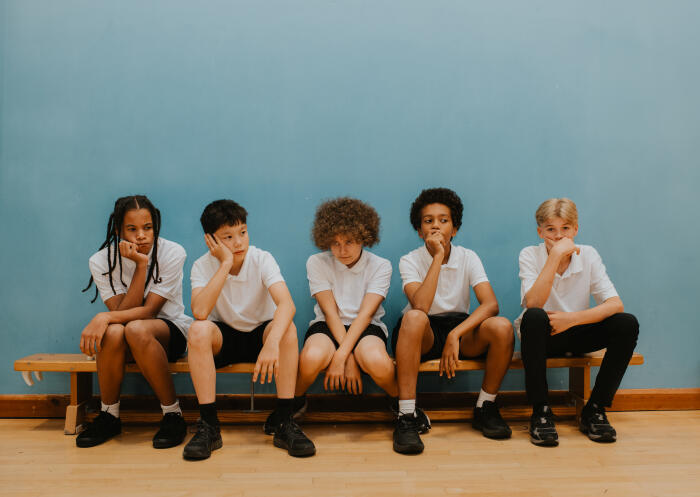 Five young teenage boys sit on a bench in a school gymnasium, looking very glum and bored.