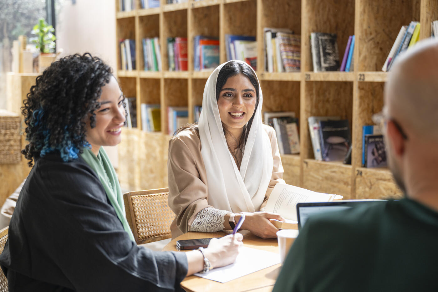 Over the shoulder view of smiling students in traditional clothing sitting at table and pausing from studying to converse.