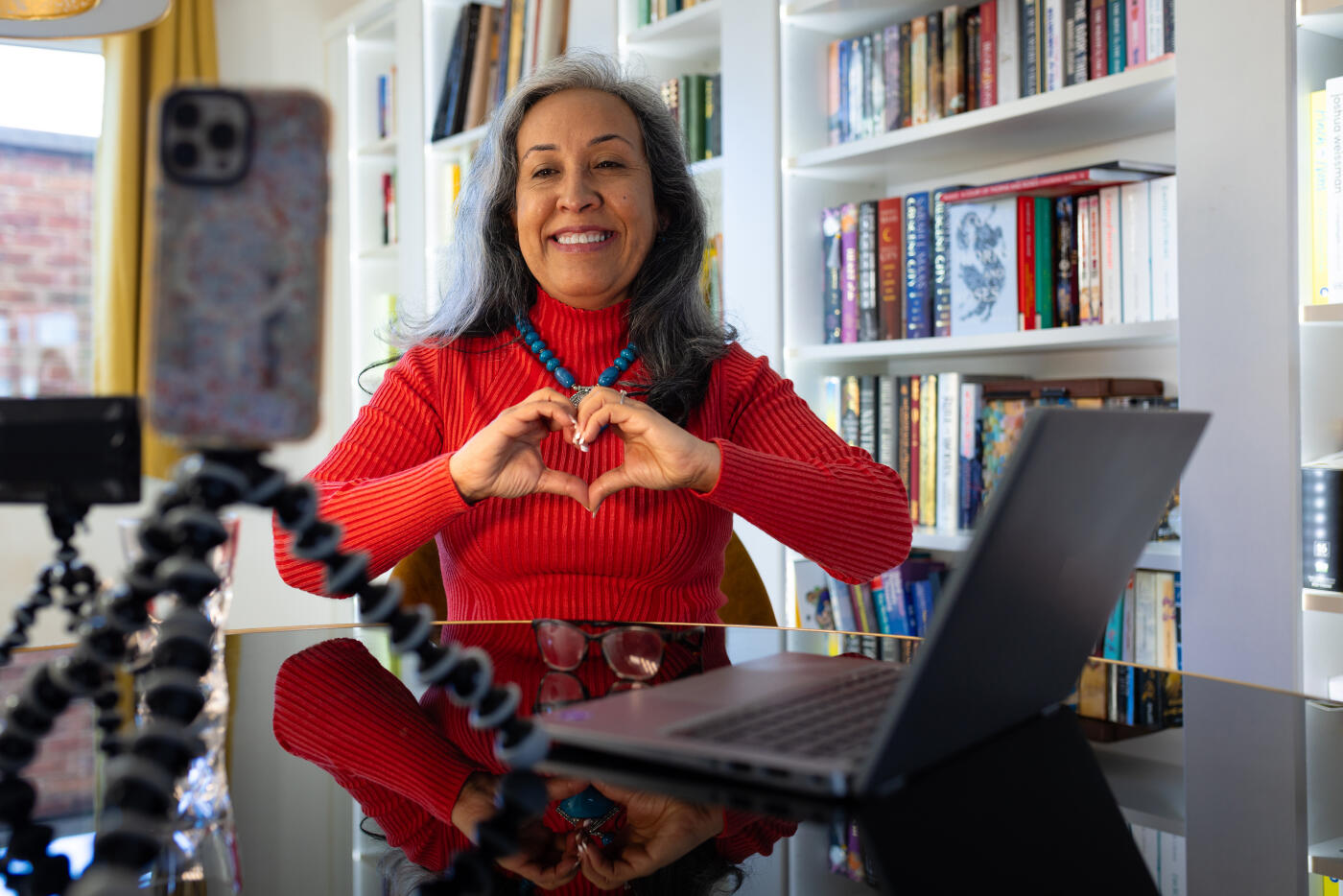 Medium shot of a mature woman sitting down at her dining table in her home in the North East of England. She has her laptop open and is using her mobile phone on a tripod to record herself as she is an influencer. The woman is looking at her phone and smiling while making a heart shape with her hands. A small light is shining on her to complete the set-up.Videos are available similar to this scenario.