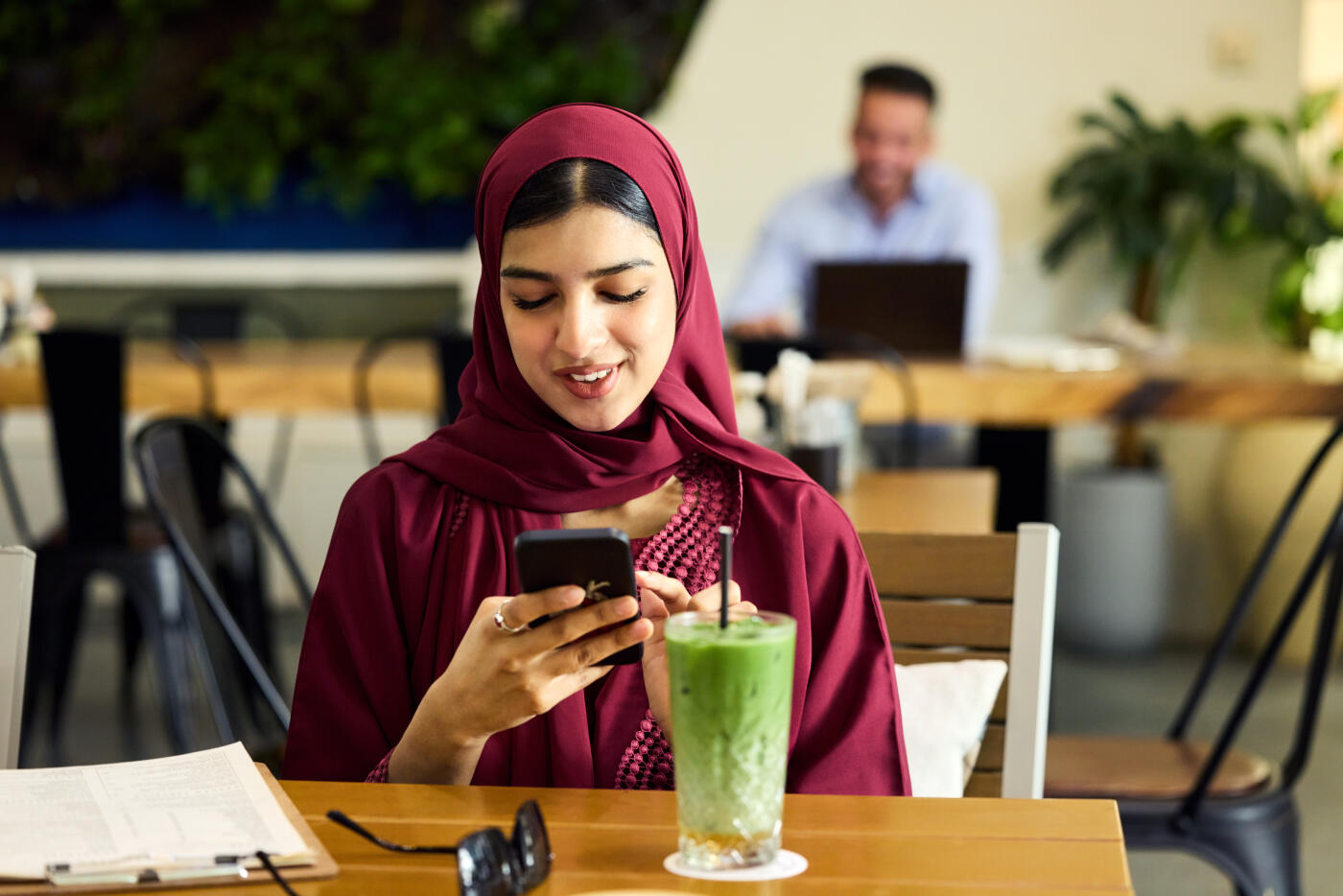 Young woman surfing through smart phone while sitting in cafe. Female is wearing hijab and abaya. Iced macha latte is kept on table.