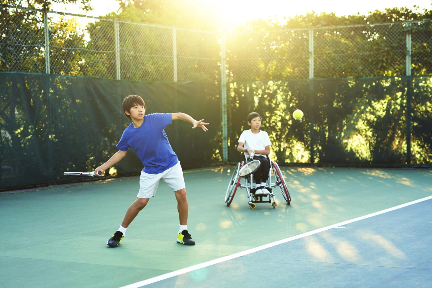 Two teenage athletes, one adaptive athlete, on an outdoor tennis court on a sunny afternoon