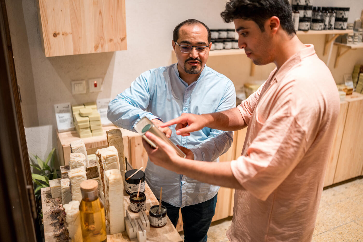 Two men, one in blue and the other in pink, explore a traditional soap shop in Al Balad, examining natural handcrafted soaps and oils in a cozy wooden interior.