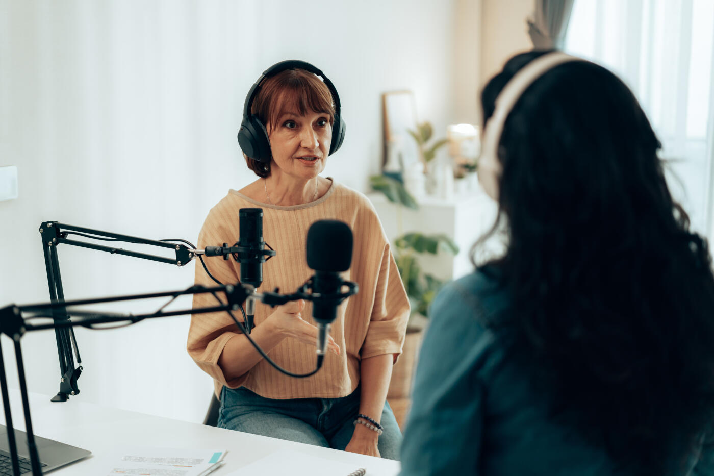SEnior Woman wearing headphones speaking into a microphone while interviewing a guest in a podcast studio. Perfect for themes of media, broadcasting, and technology.
