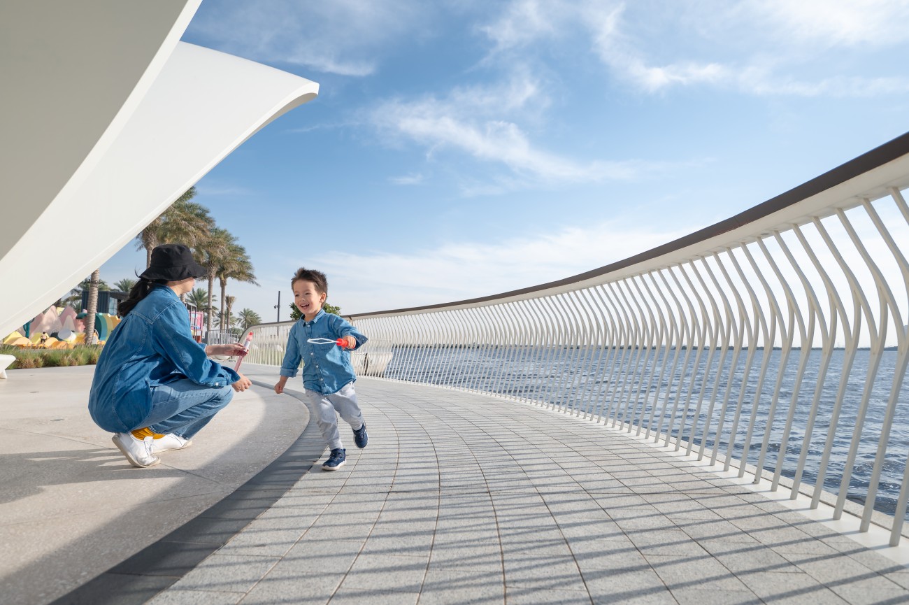 A mother and her toddler create soap bubbles with a bubble wand along the seaside promenade, enjoying a playful and joyous moment together by the coast of Dubai creek harbor public promenade