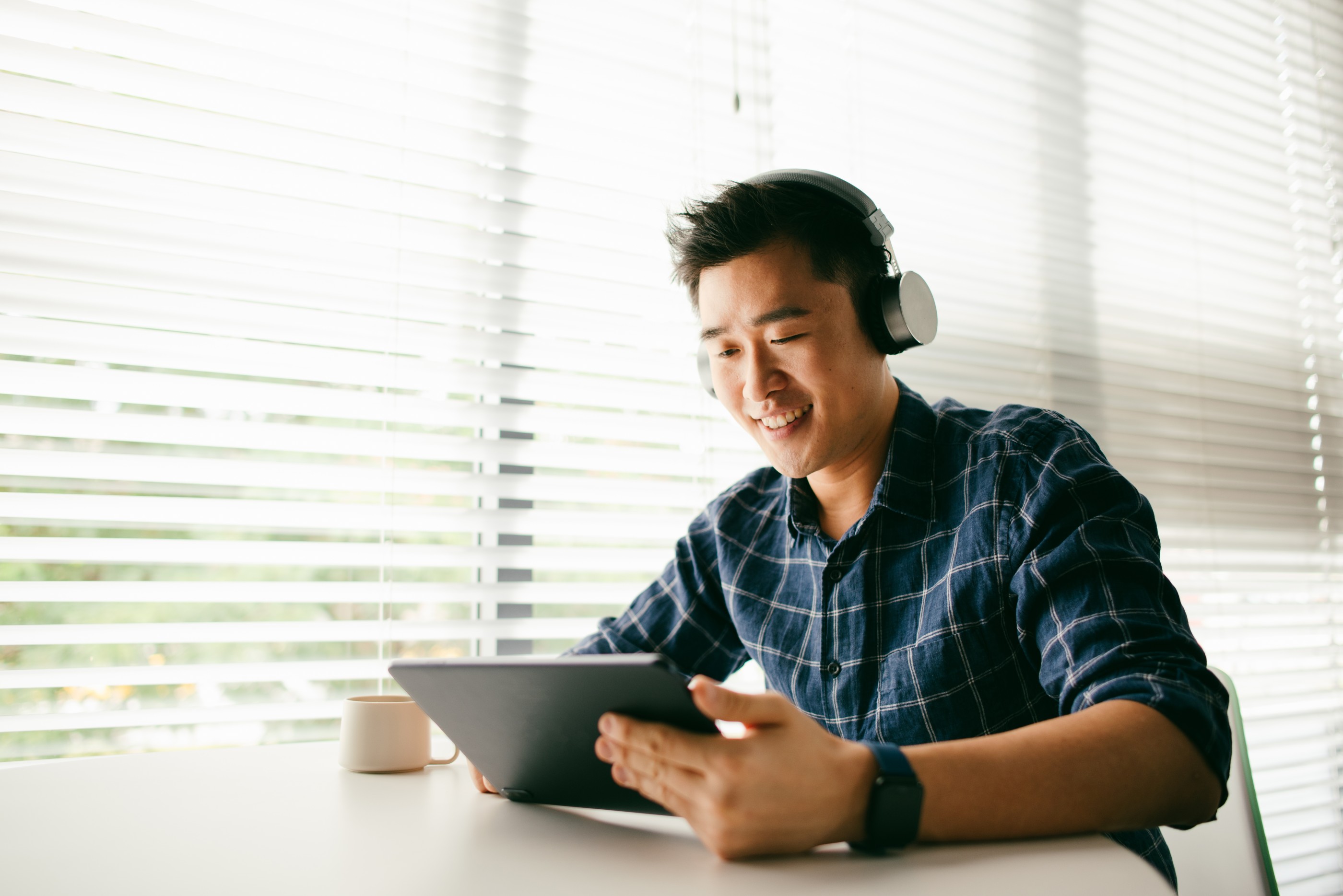 An Asian man relaxing by the home window and typing a message on his digital tablet