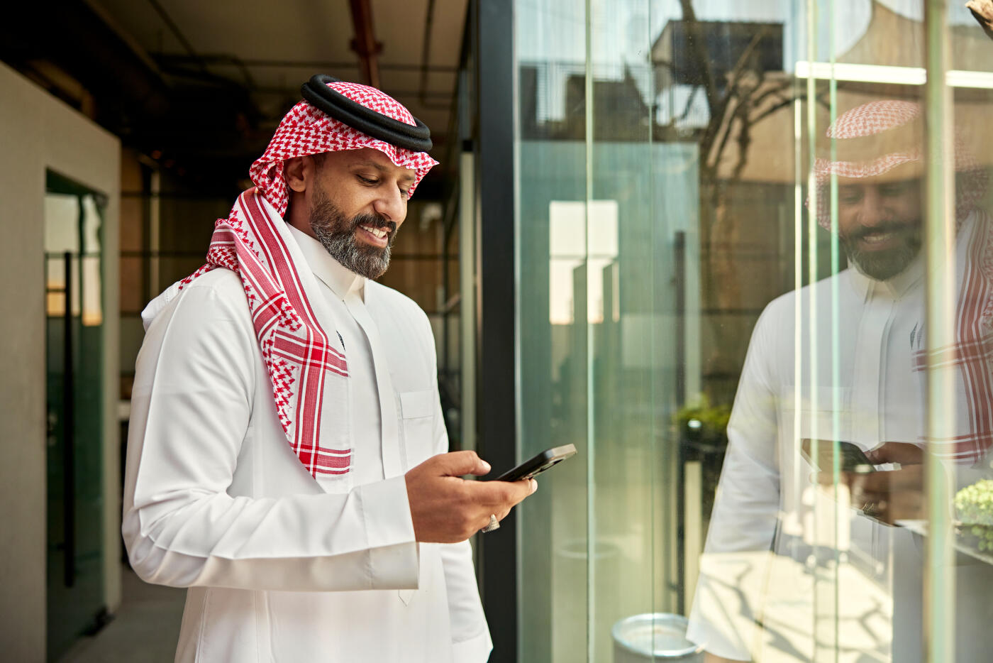 Three-quarter front view of bearded businessman in early 40s pausing in corridor of modern office to look at device.