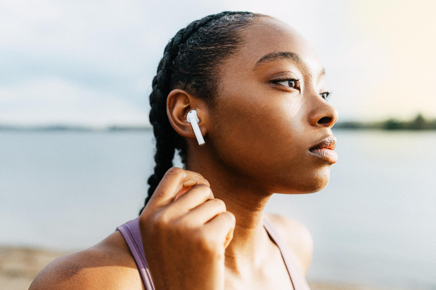 A young woman with braided hair is adjusting her wireless earbuds while outdoors near a body of water. The serene setting suggests she might be enjoying music or a podcast in a tranquil environment.