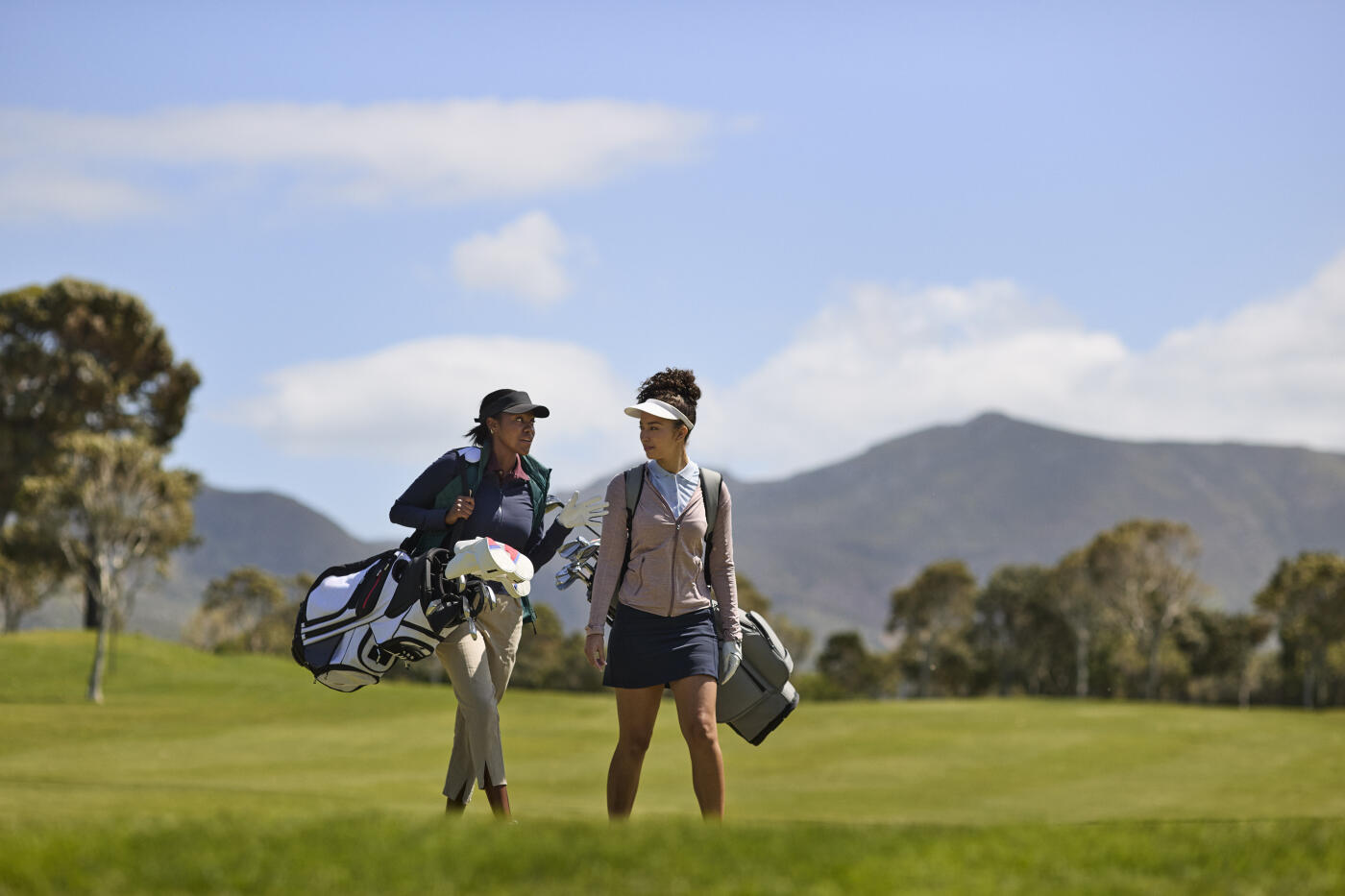 Young woman carrying golf bag talking with friend while walking on course at sunny day