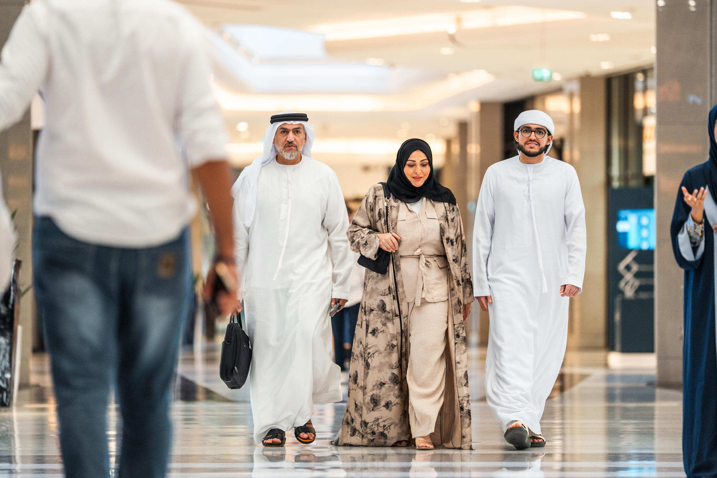 Emirati woman wearing traditional abaya and headscarf walks through sleek mall corridor flanked by two men in white thobes. Polished floors reflect warm overhead lighting. Other shoppers visible in background.